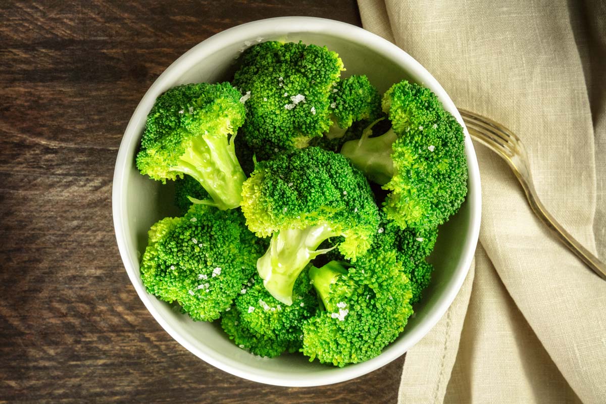 Un haut en bas d'un petit bol en céramique blanc contenant des fleurons de brocoli légèrement cuits, saupoudrés de sel de mer, avec une serviette en tissu à droite du cadre et une fourche en métal, sur une surface en bois.