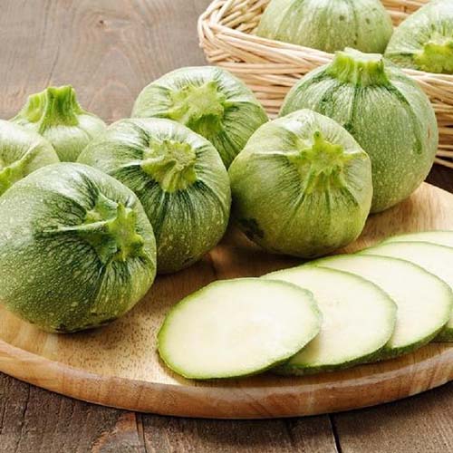 Un gros plan d'une planche à découper en bois avec une courge de courgette ronde, un peu entier et une tranchée en rondes. La variété est vert clair avec des taches légères et est appelée «Ronde de Nice». En arrière-plan est un panier en osier.