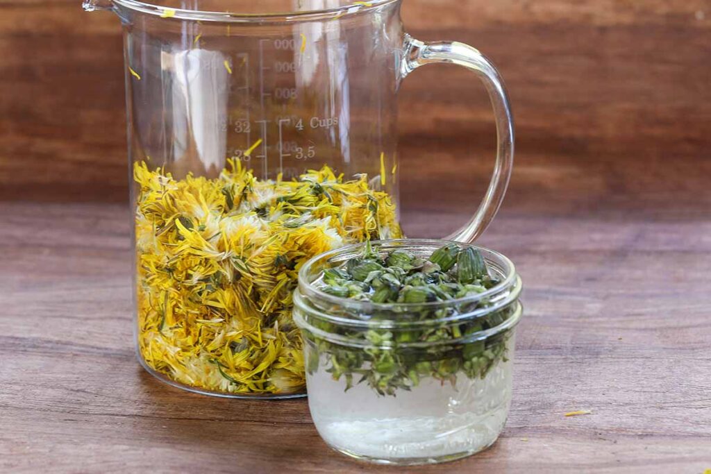Une image horizontale en gros plan de câpres de pissenlit dans un pot et de fleurs dans une cruche posée sur une surface en bois.