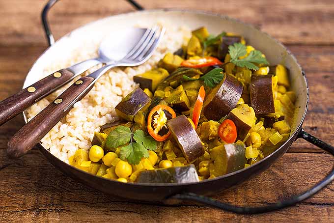 Un gros plan d'un bol en métal avec un curry d'aubergines fraîches garni de tranches de piments rouges et de coriandre, avec du riz sur le côté et une fourchette et une cuillère, avec une surface en bois en arrière-plan.